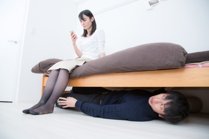 ベッドの下に潜り、ベッドに座る女性を見ている男性