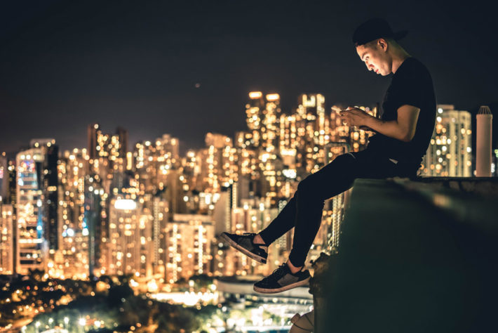 高層ビルの屋上に座ってスマホをいじっている男性